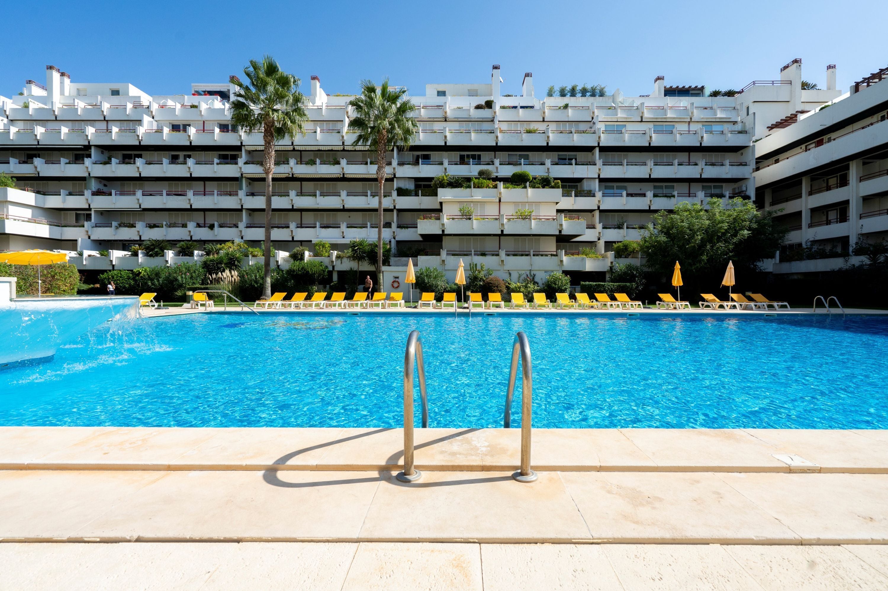 A swimming pool with a view of a multi-story building with balconies and palm trees.