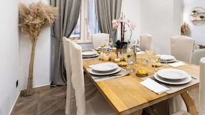 Uma sala de jantar com uma mesa de madeira posta para quatro, cadeiras brancas, um vaso com flores e uma janela com cortinas.