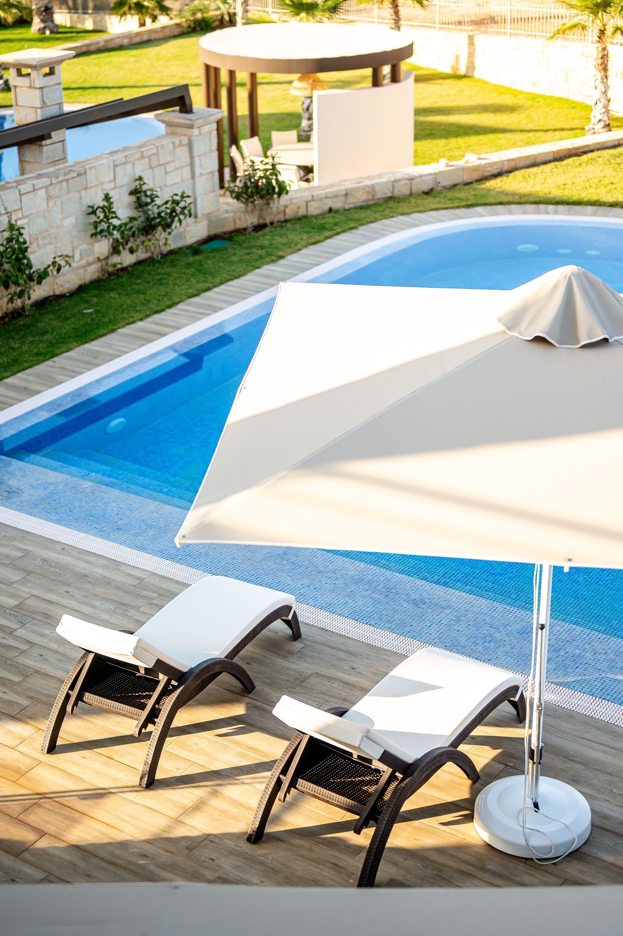 A poolside area with a covered umbrella, two lounge chairs, and a stone wall.
