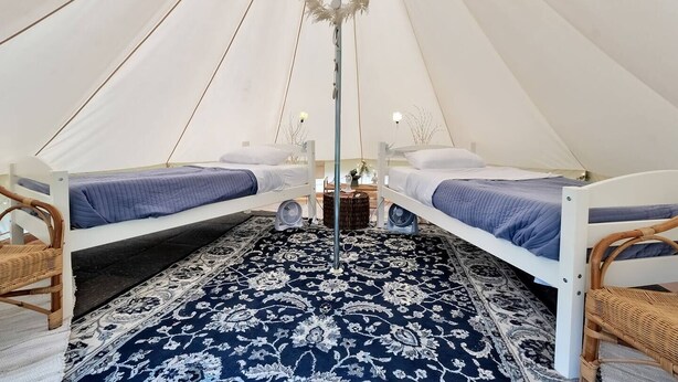 A tent with two beds, each with a blue blanket and white pillows. There is a decorative plant hanging from the ceiling and a woven basket on the floor.