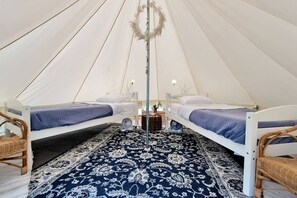 A tent with two beds, each with a blue blanket and white pillows. There is a decorative plant hanging from the ceiling and a woven basket on the floor.