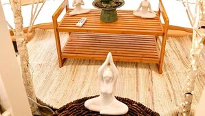 A meditation area with a wooden bench, two white statues, a small table with a green object, and a woven rug.