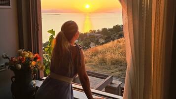 A room with a large window overlooking a sunset over a body of water, a potted plant, and a piano.