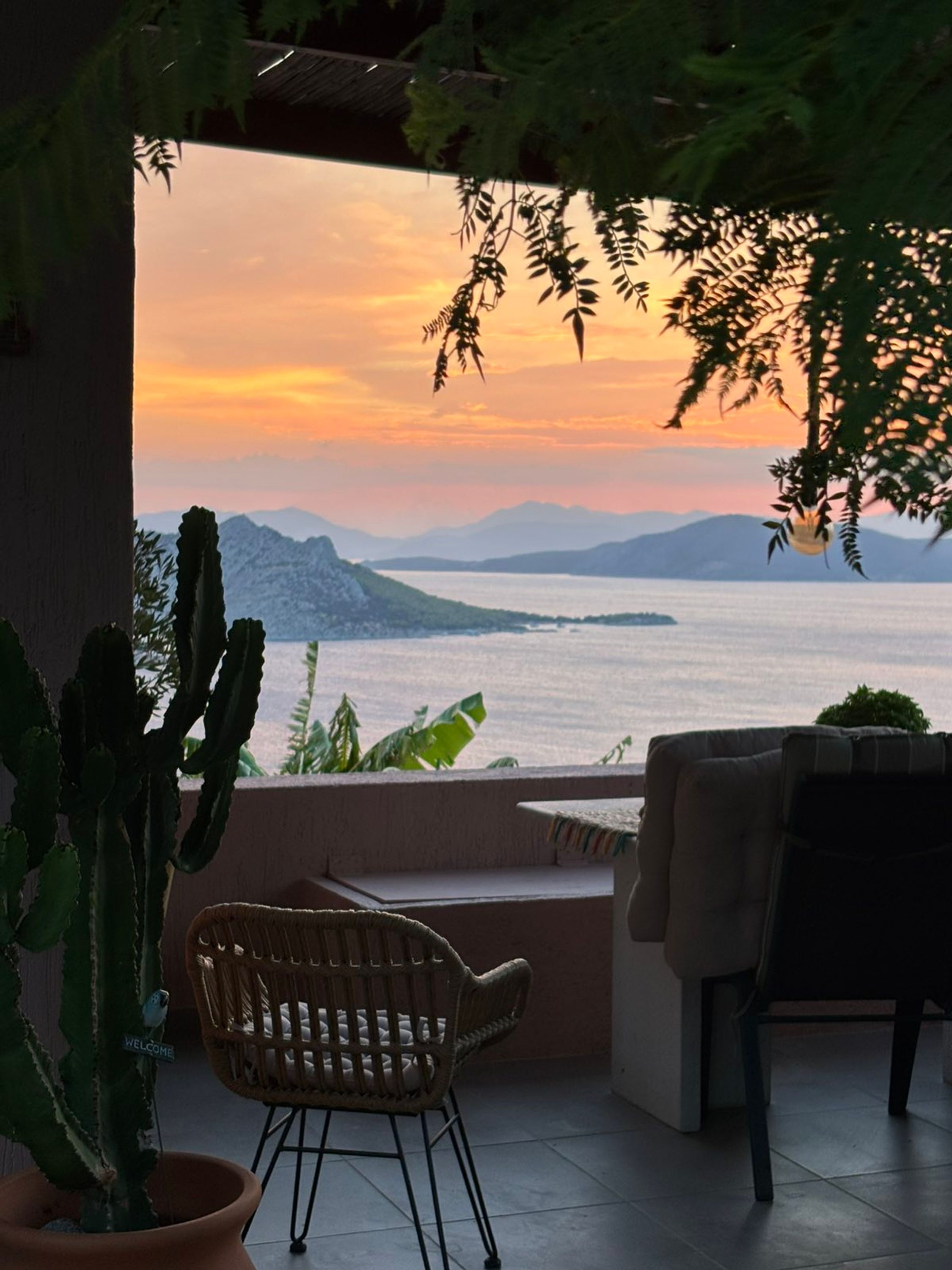 A patio with a view of a sunset over a body of water and mountains, a cactus plant, and a wicker chair.