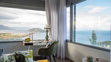 A room with a large window offering a view of a coastal landscape, a desk with a lamp, a chair, and decorative pillows.