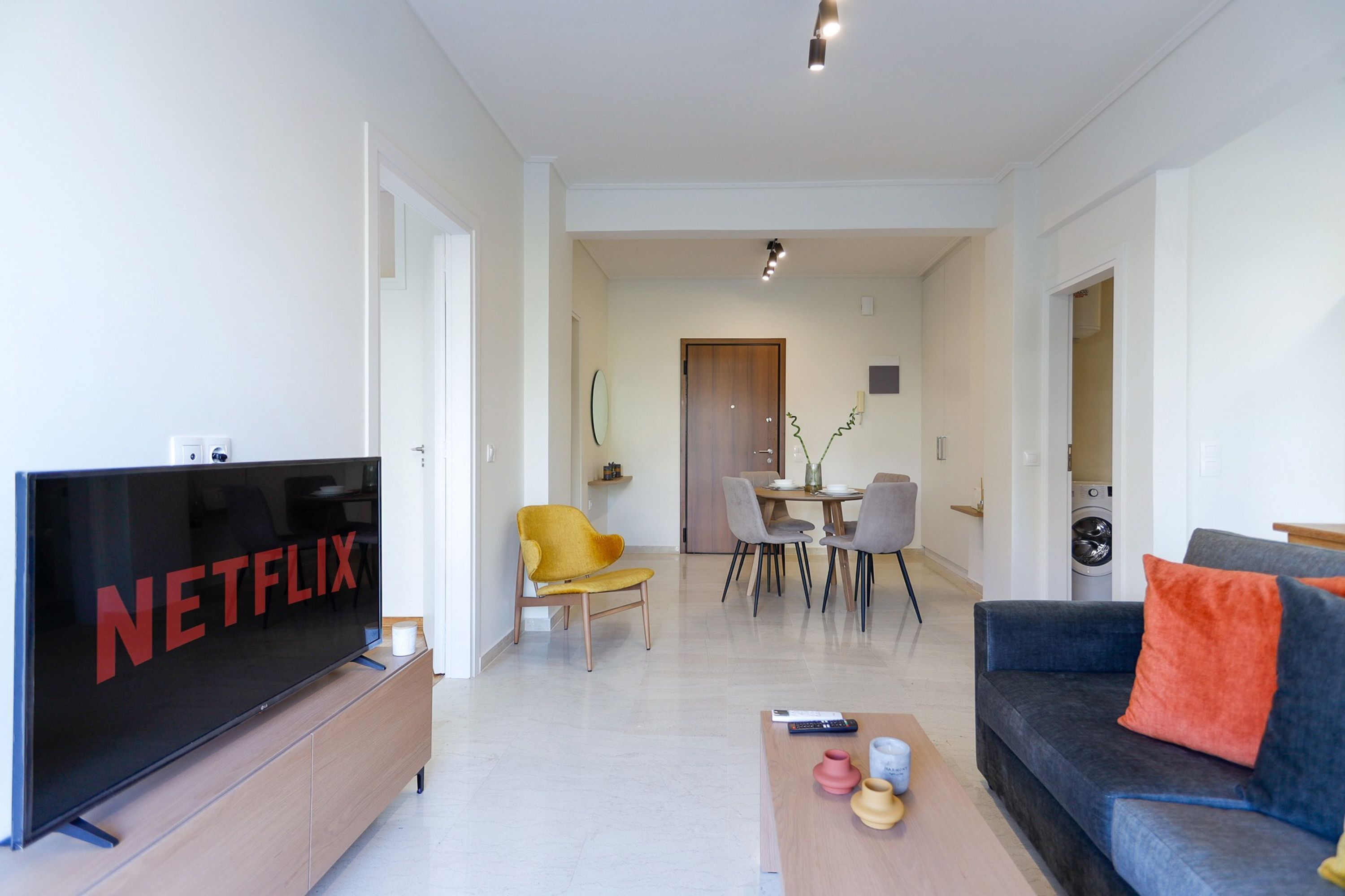 A modern living room with a flat-screen TV, a sofa, a coffee table, and a dining area with chairs and a table.