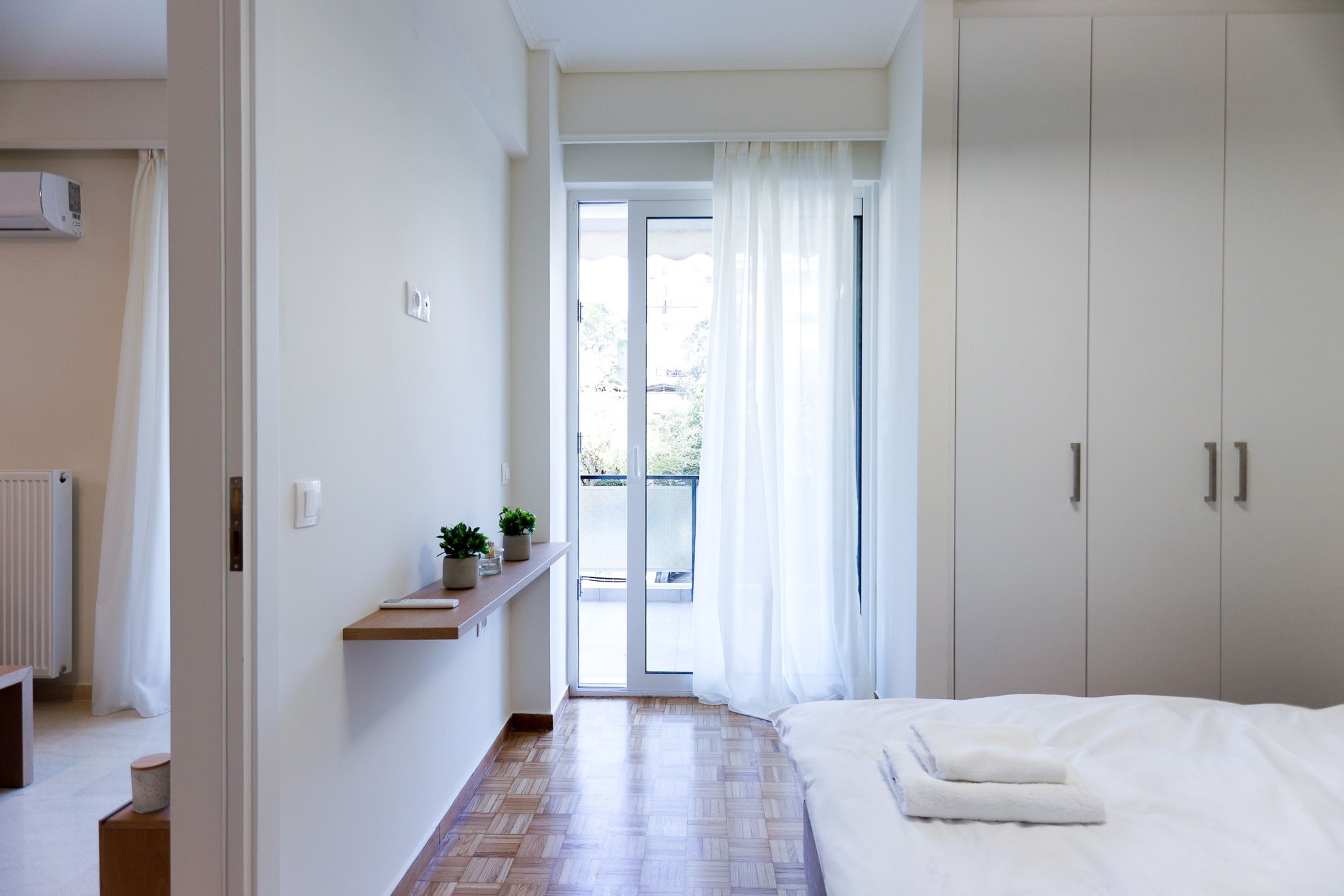 A bedroom with a bed, a wardrobe, a radiator, a shelf with a plant, and a view of the outside.