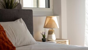 Un lit bien fait, avec une literie blanche, une tête de lit grise, une petite plante en pot aux fleurs violettes et une lampe sur une table de chevet.
