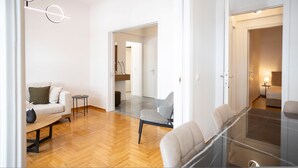 Un salon moderne avec un canapé, une table basse en verre et un fauteuil. Un couloir mène à une autre pièce où se trouve une lampe.