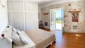 A bedroom with a bed, a chair, a painting, and a view through a glass door. - Villa Joy in Ampelas, Paros (Ampelas)