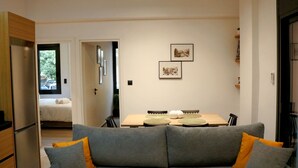 A modern living room with a grey sofa, a wooden dining table, and a kitchen area with a refrigerator. - No 2 - Theo Apartment two bedrooms ground floor (Athina)