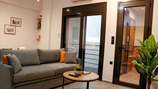 A modern living room with a grey sofa, a small round table with a plant, and a sliding glass door leading to a balcony. - No 2 - Theo Apartment two bedrooms ground floor (Athina)