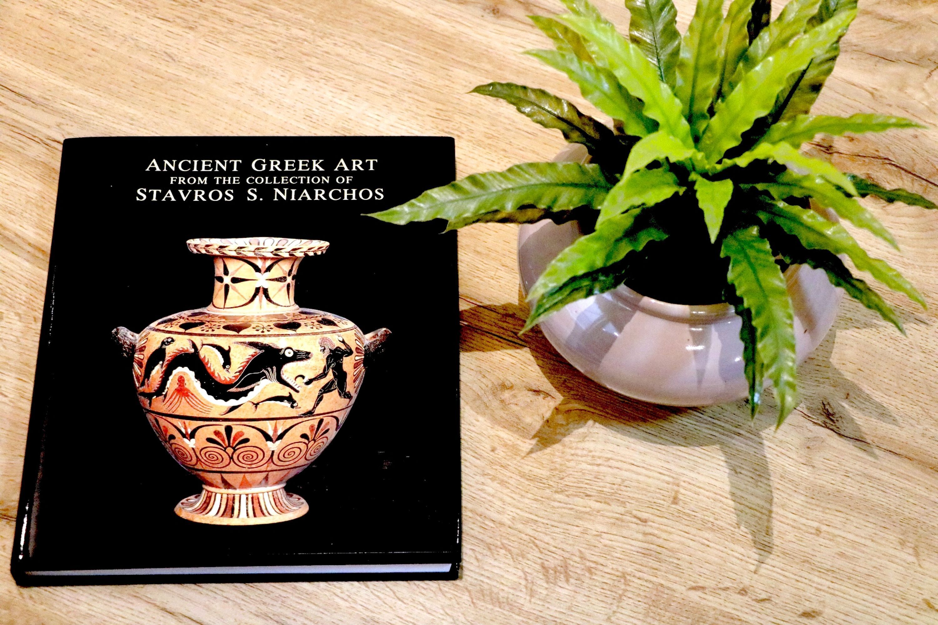 Un libro titulado "Arte Griego Antiguo de la Colección Stavros S. Niarchos" sobre una superficie de madera, junto a una planta en maceta.