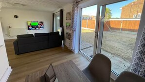 Un salon avec un canapé noir, un téléviseur fixé au mur et une porte-fenêtre coulissante donnant sur un jardin.