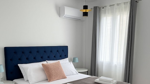 Ein ordentlich bezogenes Bett mit blauem Kopfteil, weißer Bettwäsche und einer braunen Tagesdecke. Ein kleiner Nachttisch mit Lampe steht daneben, und ein Fenster mit Vorhängen ist ebenfalls zu sehen.
