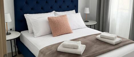 Ett modernt hotellrum med en stor säng, ett stoppat sänggavell, vita sängkläder, en brun pläd och ett litet nattduksbord med en lampa.