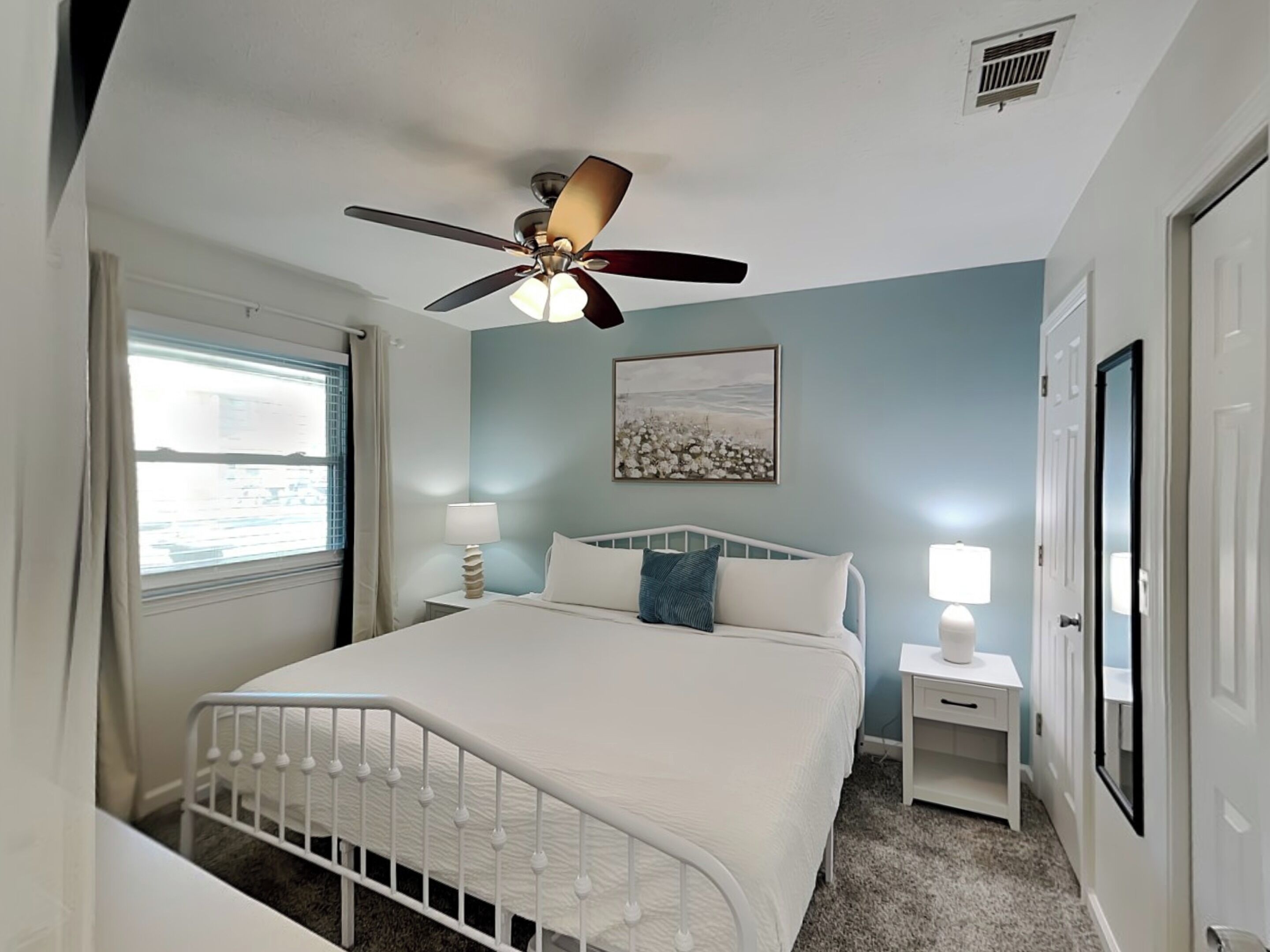 Une chambre à coucher avec un lit, un ventilateur de plafond, une fenêtre avec des rideaux, une table de chevet avec une lampe et un tableau accroché au mur.