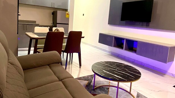A modern living room with a sofa, a dining table with chairs, a ceiling fan, and a flat-screen TV mounted on the wall. - Two Bedroom Apartment, BH Apartments Dansoman Heights (Dansoman)