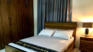 A bedroom with a bed, a ceiling fan, a nightstand, a wardrobe, and a bench. - Two Bedroom Apartment, BH Apartments Dansoman Heights (Dansoman)