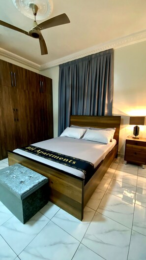 A bedroom with a bed, a ceiling fan, a nightstand, a wardrobe, and a bench. - Deluxe One Bed room Apartment, BH Apartments (Dansoman)