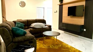 A living room with a sofa, a coffee table, a TV mounted on the wall, and a ceiling fan. - Two Bedroom Apartment, BH Apartments Dansoman Heights (Dansoman)