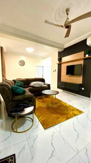 A living room with a sofa, a coffee table, a TV mounted on the wall, and a ceiling fan. - Deluxe One Bed room Apartment, BH Apartments (Dansoman)