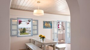 Une pièce avec une table et des bancs blancs, un luminaire suspendu et une vue par une fenêtre à volets.