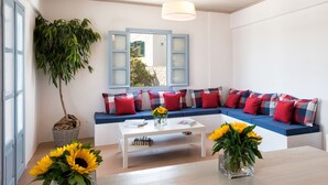 Un salon moderne avec un canapé blanc, des coussins bleus et une grande fenêtre dotée de volets bleus. On y trouve deux vases contenant des tournesols, posés sur une table basse en bois.