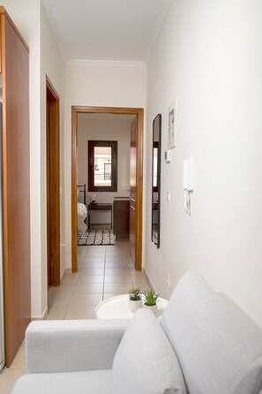 Ein Flur, der zu einem Schlafzimmer führt, mit Bett, Nachttisch und Fenster. An der weißen Wand befindet sich ein aufgehängtes Bilderrahmen und eine Wandleuchte.