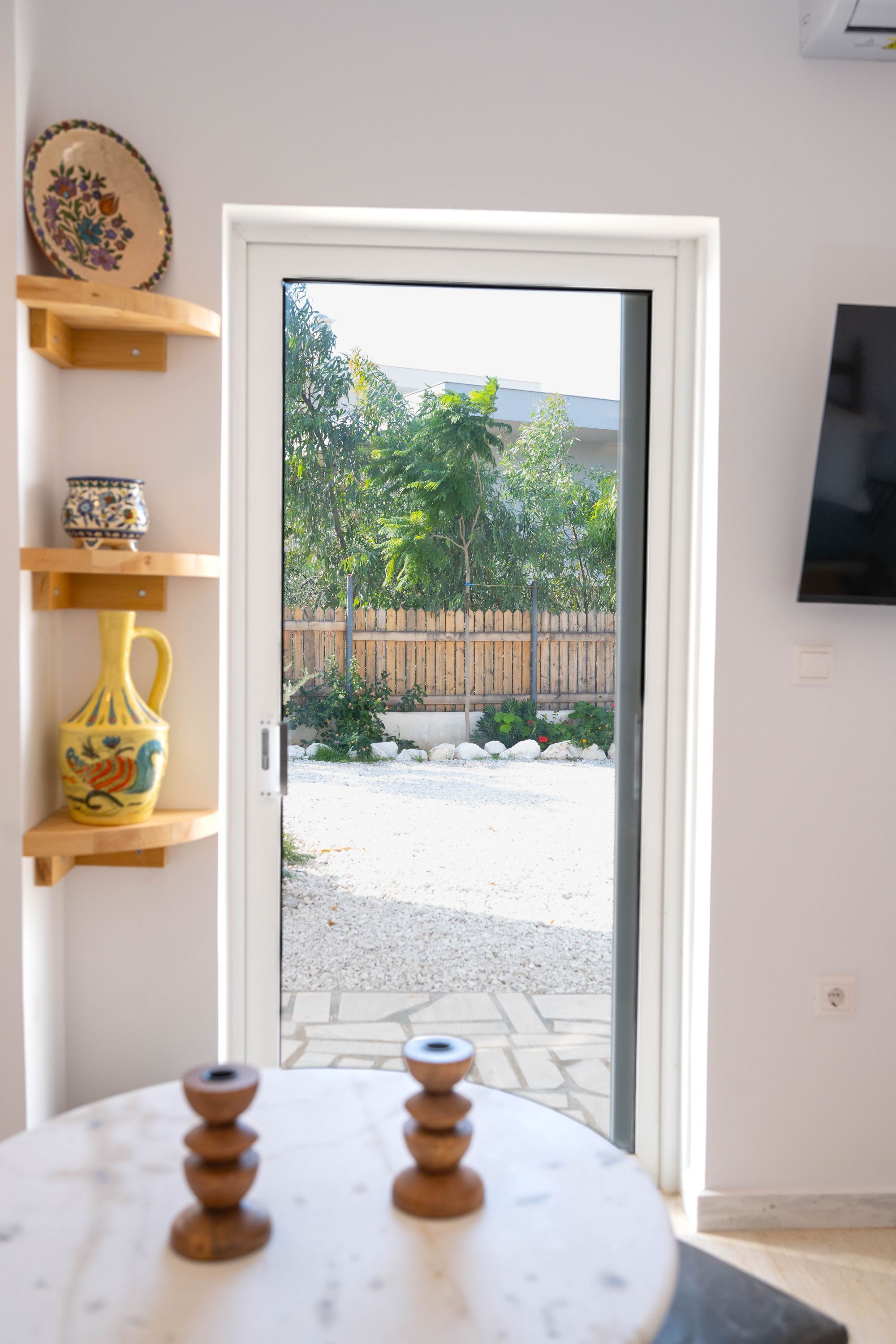 Una habitación con puerta que da a un jardín, un televisor montado en la pared y estanterías con adornos.