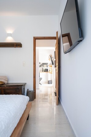 Une chambre à coucher comprenant un lit, une table de chevet en bois, un téléviseur fixé au mur, et un couloir menant à un espace cuisine.