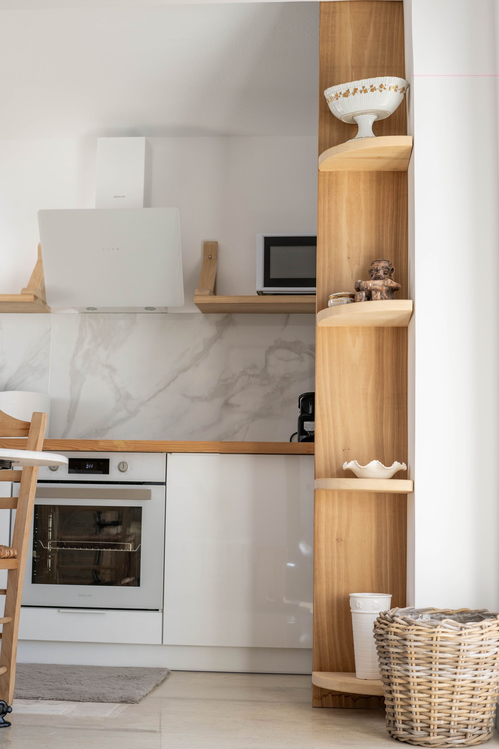 Une cuisine moderne avec des meubles blancs, un four encastré, un four à micro-ondes et une étagère en bois où sont disposés des objets décoratifs.