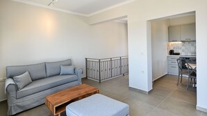 Ein modernes Wohnzimmer mit einem grauen Sofa, einem Holztisch und einem grauen Hocker. Es gibt eine Küchenzeile mit einem Esstisch und Stühlen sowie einen kleinen Balkon mit Geländer.