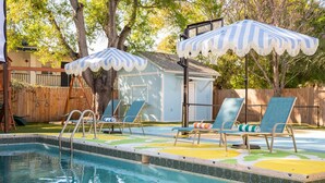 Une piscine entourée d’une clôture en bois et d’arbres, avec des chaises longues et des parasols à rayures.