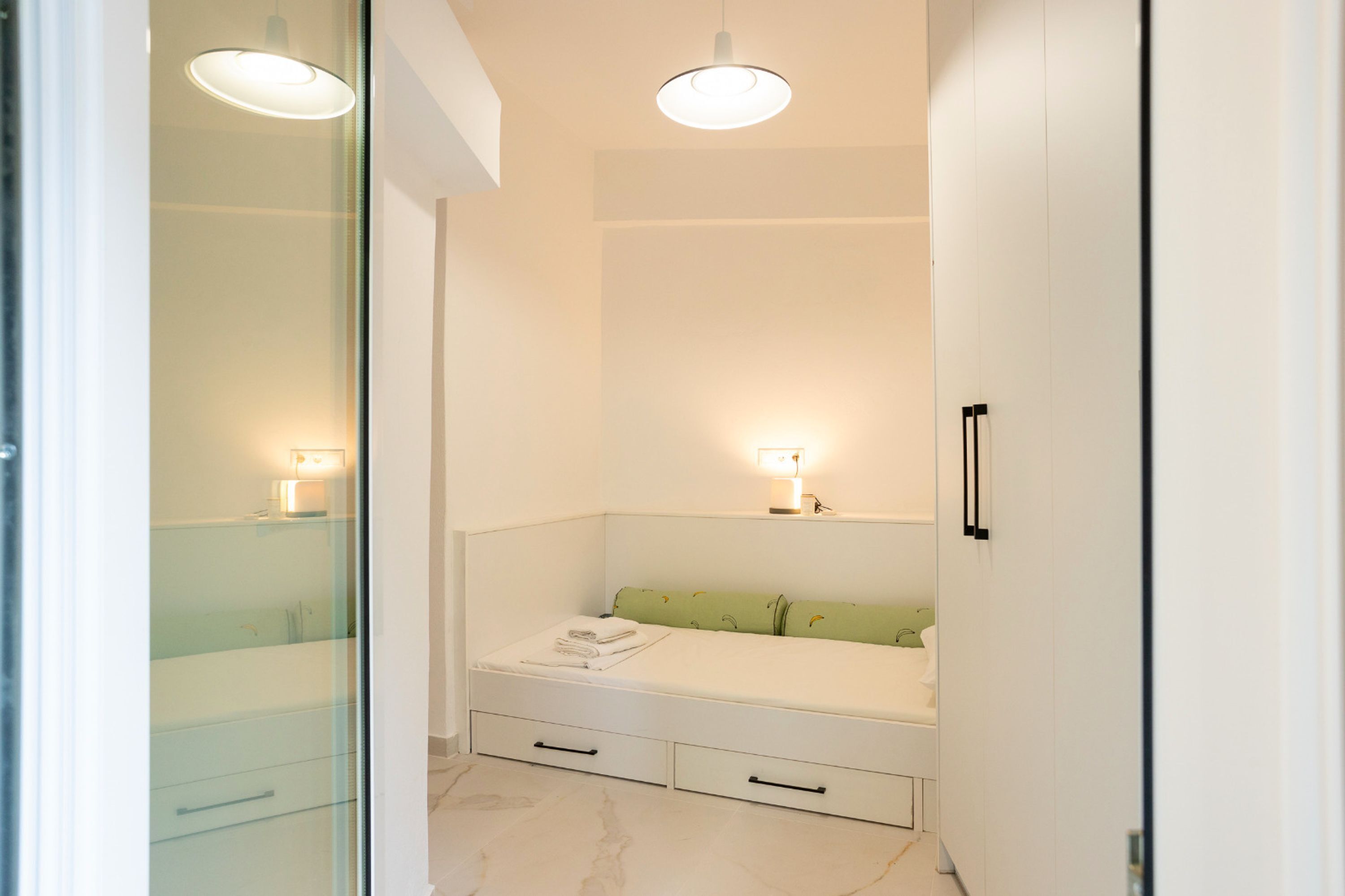 Une salle de bain moderne et minimaliste, dotée d’une douche en verre, d’un meuble-lavabo blanc avec un miroir et d’un lit avec une tête de lit verte.