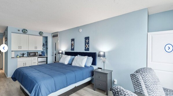 A bedroom with a bed, bedside tables, a chair, and a kitchenette. - King Studio with Partial Water Views, Heated Pool, Beach Access (Daytona Beach)