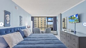 A hotel room with a large bed, a TV, a dresser, and a view of buildings through the window. - King Studio with Partial Water Views, Heated Pool, Beach Access (Daytona Beach)