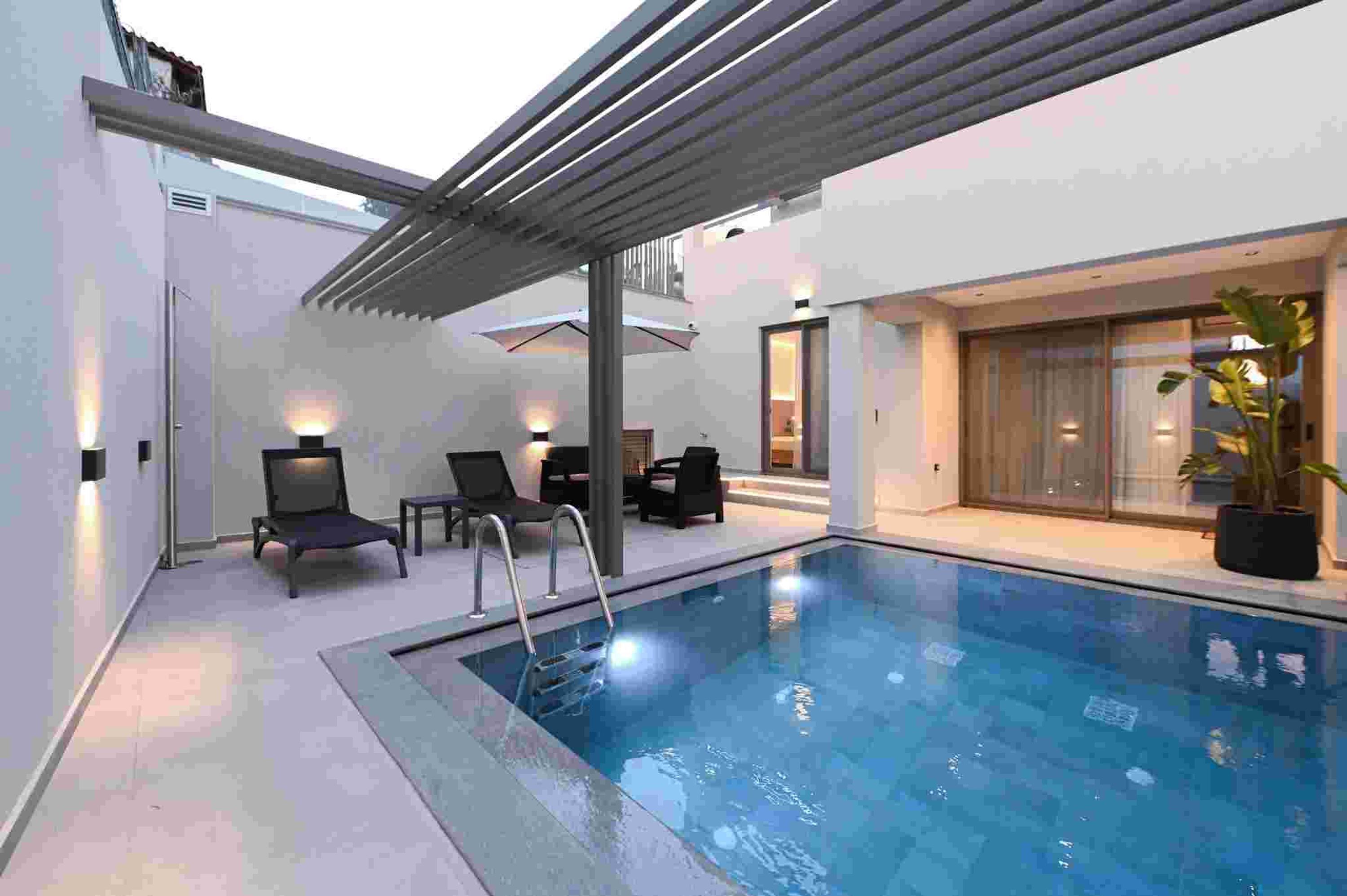 A modern indoor swimming pool with a glass enclosure, lounge chairs, and a pergola.