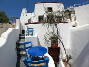 Une ruelle étroite aux murs blancs, avec une chaise et une table bleues, une porte en bois et un petit arbre.
