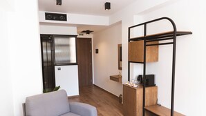 Un saló modern amb un sofà gris, un sòl de fusta, un moble prestatges de metall negre, i una zona de cuina amb nevera i microones.