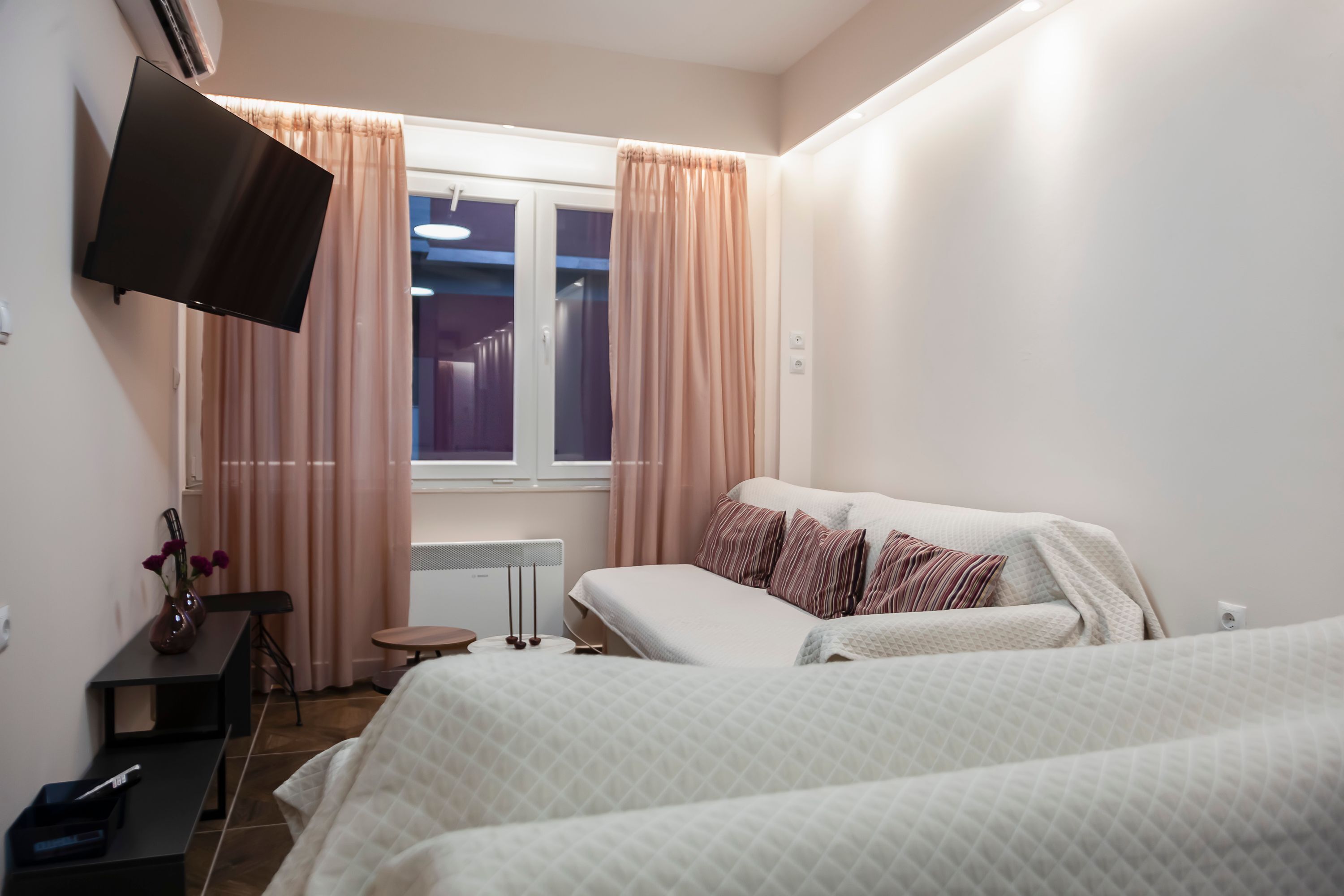 Quarto de hotel com duas camas, televisão de tela plana na parede, escrivaninha com cadeira, mesinha e janela com cortinas.