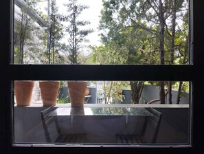 Ein Zimmer mit Glastisch und zwei Stühlen, mit Blick auf einen Garten mit Bäumen und Kübelpflanzen.