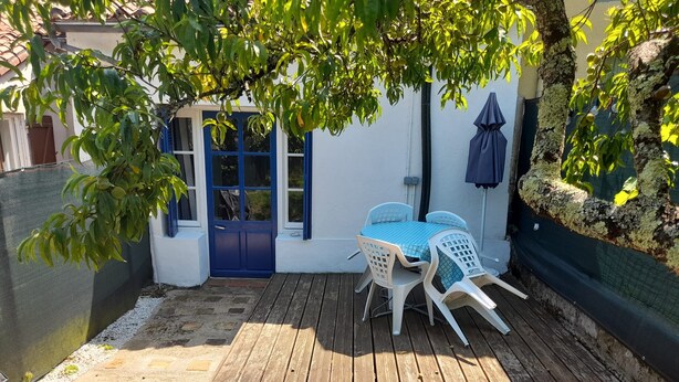 Eine Holzterrasse mit Tisch und Stühlen, umgeben von einer weißen Mauer und einer blauen Tür.