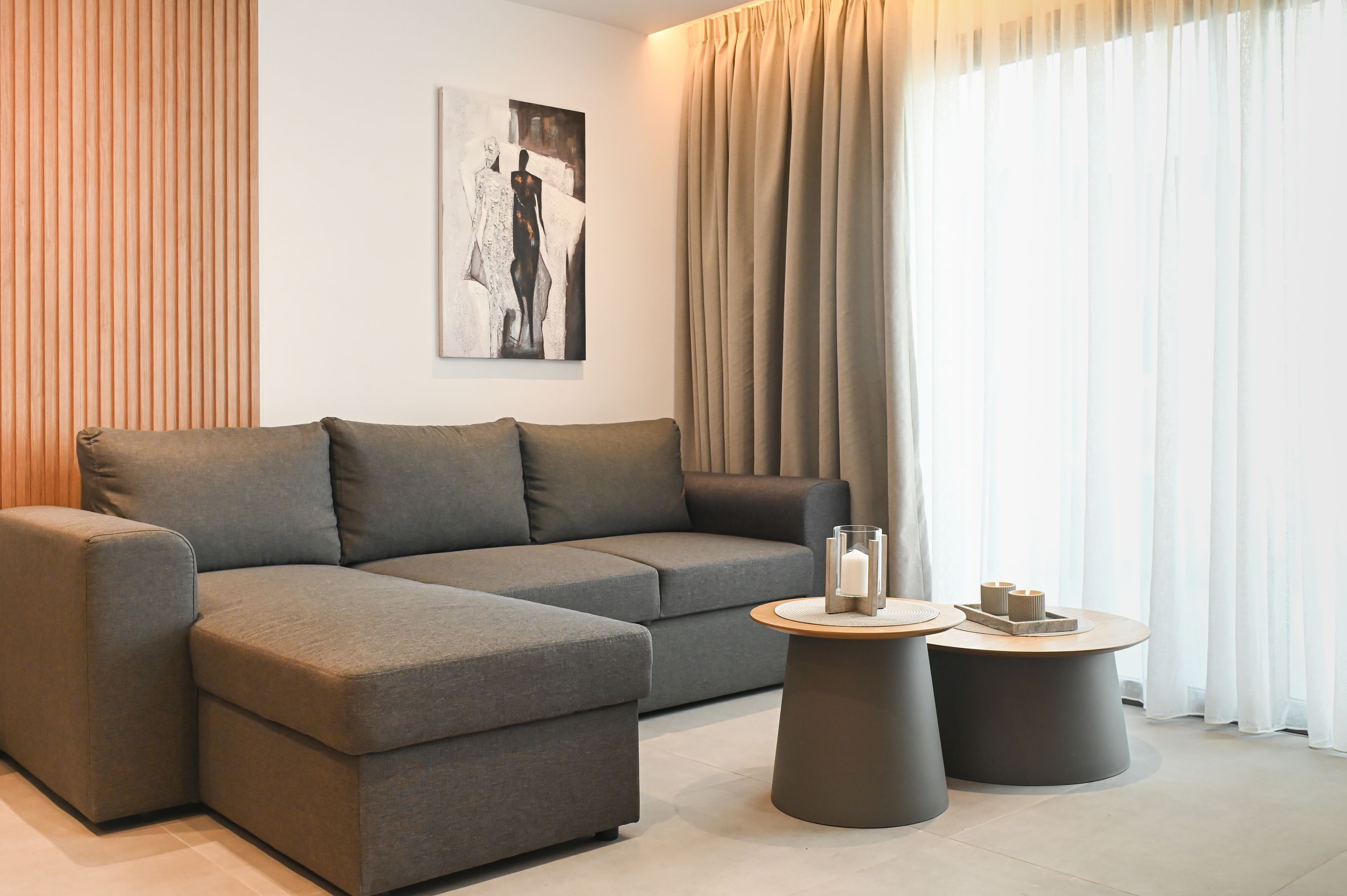 Een moderne woonkamer met een grijze modulaire bank, twee ronde salontafels en een groot raam met dunne gordijnen.