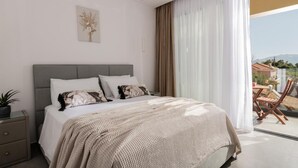 Un dormitorio con cama, mesita de noche, planta en maceta, ventana con cortinas y vistas al exterior.