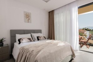 Ein Schlafzimmer mit einem Bett, einem Nachttisch, einer Zimmerpflanze, einem Fenster mit Vorhängen und Blick ins Freie.