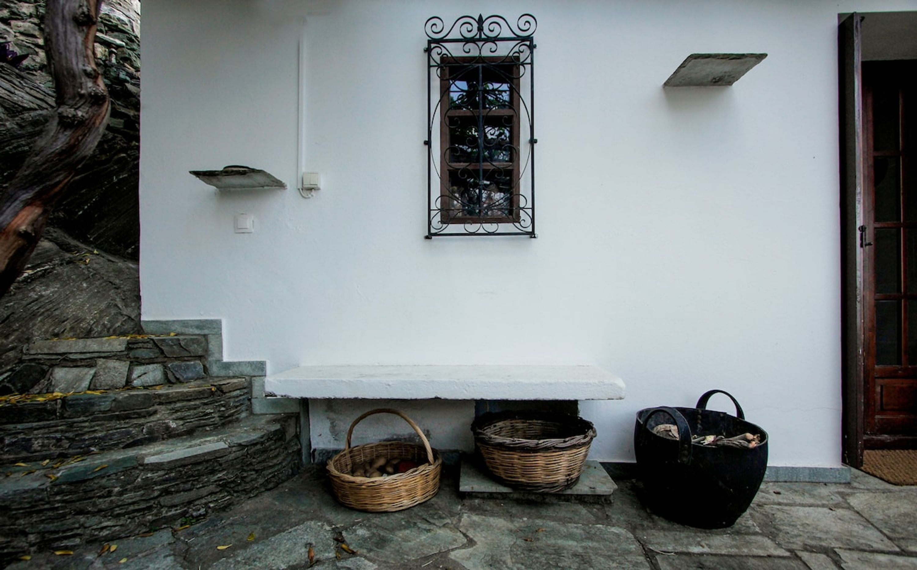 Un edificio blanco con una escalera de piedra, una ventana con rejas de hierro decorativas, un banco y dos cestas.