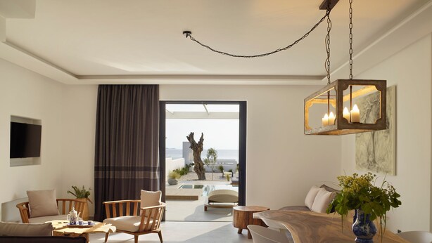 A modern living room with a large wooden dining table, comfortable seating, a TV mounted on the wall, and a view of the beach through an open door. - Two bedroom villas with private pool (Kalamitsa)