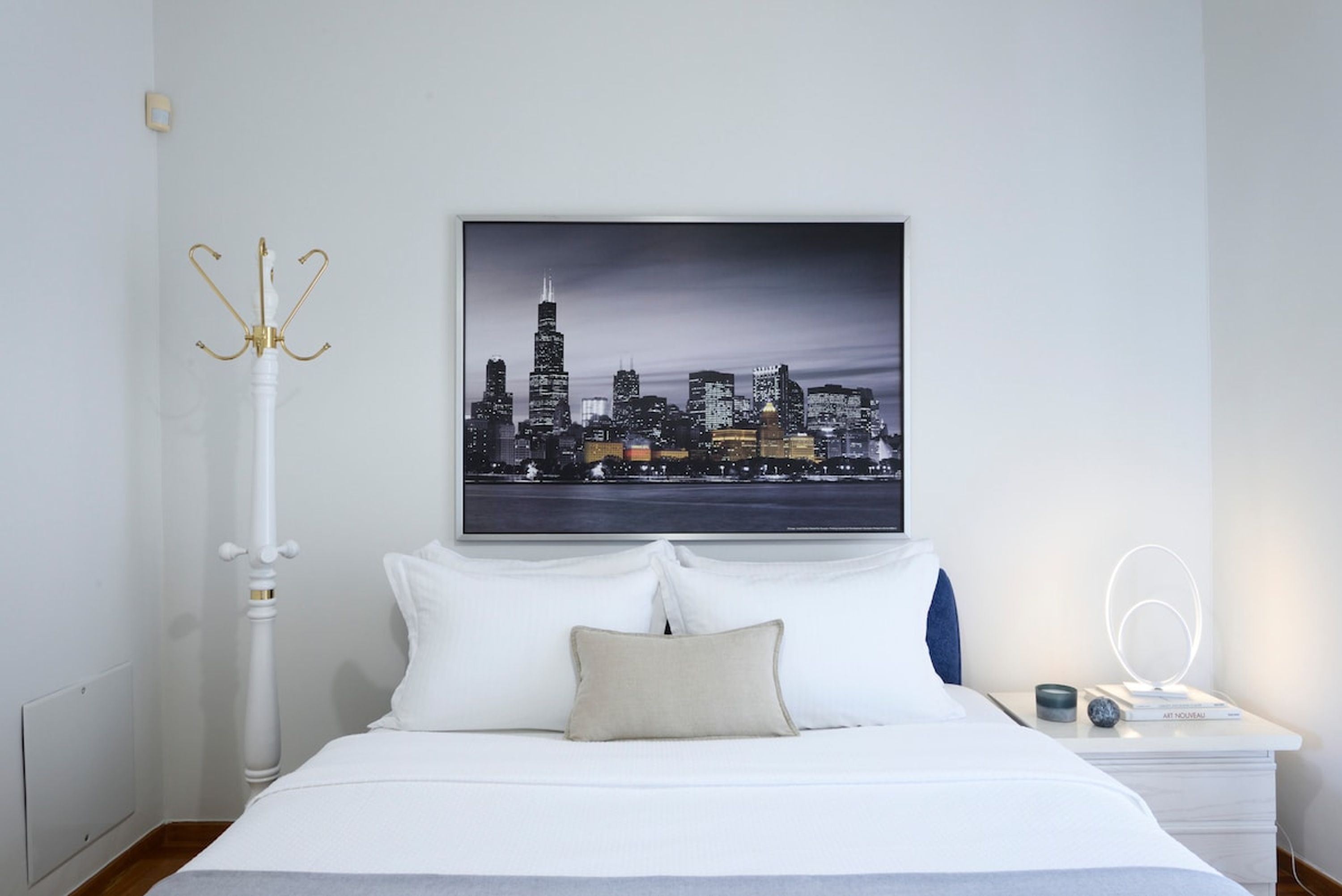 A neatly arranged bedroom with a cityscape view, a white bed, a bedside table with a lamp, and a wall-mounted light fixture.