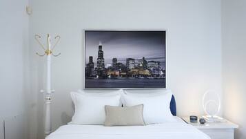 A neatly arranged bedroom with a cityscape view, a white bed, a bedside table with a lamp, and a wall-mounted light fixture.
